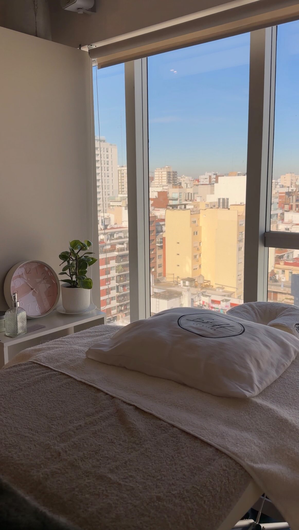 Una cabina de ByH Estética: la camilla con toallas blancas y la almohada con el logo bordado, frente a un ventanal con vista a la ciudad